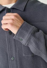 Grau gestreifte Cord-Jacke mit Kragen, geripptem Textur und grünen Druckknöpfen an den Bündchen und vorne. Schlichte Taschendetails.