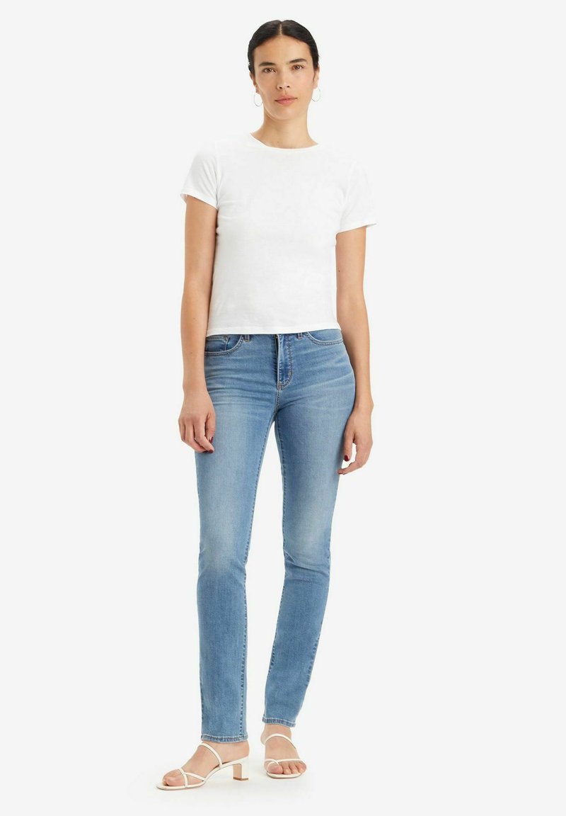 Camiseta blanca de manga corta combinada con jeans ajustados de color azul claro y sandalias blancas con tiras. La modelo está de pie con los brazos relajados a los lados.