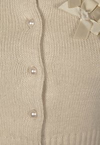 Beige stickad cardigan med pärlknappar och en mjuk textur, med en accent av krämfärgad banddetalj nära halsringningen.