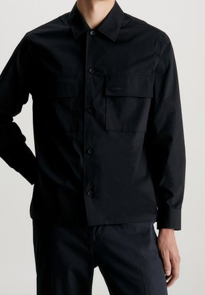 Mann trägt ein schwarzes Hemd mit Knöpfen, zwei vorderen Klappentaschen und langen Ärmeln, kombiniert mit dunkler Hose, vor einfarbigem Hintergrund.