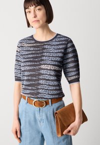 Strikket kortermet genser i mørkegrått med blå og kremfargede bladmønstre, kombinert med lyseblå jeans, brun semsket skinnbelte og clutch.
