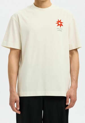 Mężczyzna ubrany w białą koszulkę z krótkim rękawem i okrągłym dekoltem, z małą czerwoną gwiazdą i napisem „Nous vous selected” na lewej piersi.