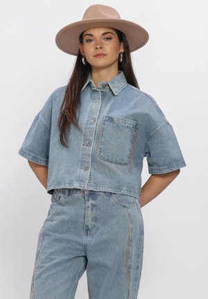 Vrouw draagt lichtblauw denim cropped overhemd en hooggetailleerde jeans met zilveren hoepeloorbellen en beige hoed met brede rand, staand tegen een witte achtergrond.
