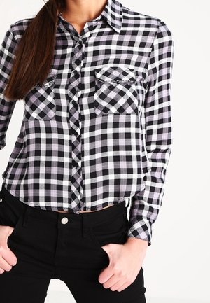 Black and white checkered button-up shirt with chest pockets worn with black pants, hands tucked into front pockets.
