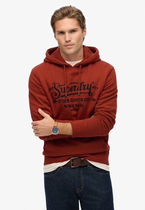 Rode hoodie met een opvallend zwart "Superdry" logo, koordjes en geribbelde boorden, gecombineerd met donkere jeans en een bruine horloge.