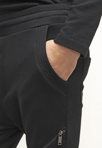 Černé joggingové kalhoty vyrobené z měkké tkaniny, s bočními kapsami na zip a žebrovaným detailem na pase. Texturovaný a přiléhavý design.