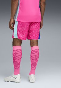 Różowa koszulka sportowa z krótkim rękawem z akcentami w kolorze aqua, w towarzystwie różowych spodenek we wzory i różowych skarpet do kolan, noszona z białymi butami sportowymi.