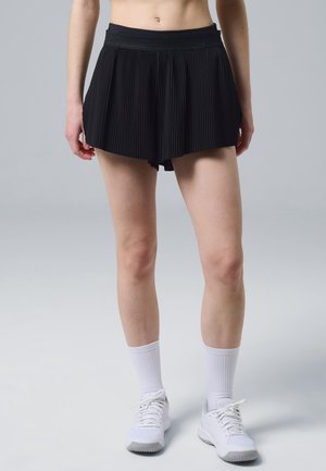 Persona que lleva una falda deportiva negra plisada, calcetines blancos hasta la pantorrilla y zapatillas blancas, de pie sobre un fondo gris.