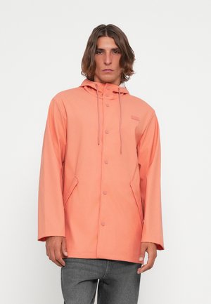 Peach-colored, knee-length rain jacket with a hood, snap buttons, side pockets, and a relaxed fit. Smooth, lightweight fabric.