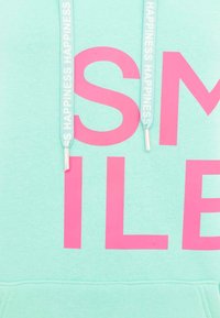 Mint green hoodie featuring large pink text that reads "SMILE." White drawstrings with repeating text pattern, smooth texture.