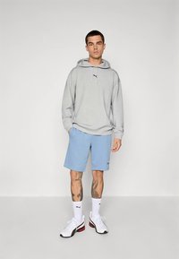 Hanorac gri deschis cu buzunar frontal tip sac și logo roșu, combinat cu pantaloni scurți albaștri și adidași albi cu accente roșii. Tatuaje vizibile pe picior.