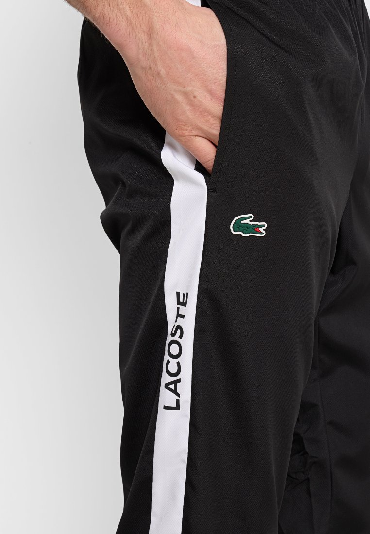 Pantalons de sport noirs en tissu texturé. Présente une rayure blanche et un détail du logo Lacoste sur le côté. Taille élastique pour le confort.