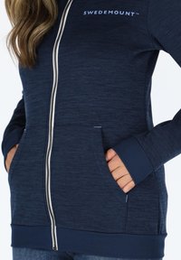 Felpa con zip di colore blu navy, con texture a maglia. Presenta due tasche laterali, polsini a costine e un logo sul petto. Zip argentata.