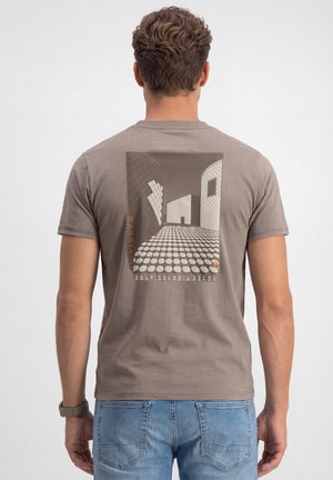 Man draagt een grijs T-shirt met architectonisch ontwerp met gebogen muren en een gestippelde vloer, gecombineerd met lichtblauwe jeans.