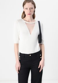 Un haut en tricot blanc avec un décolleté en V profond et des manches courtes, associé à un pantalon noir avec des boutons dorés sur le côté.