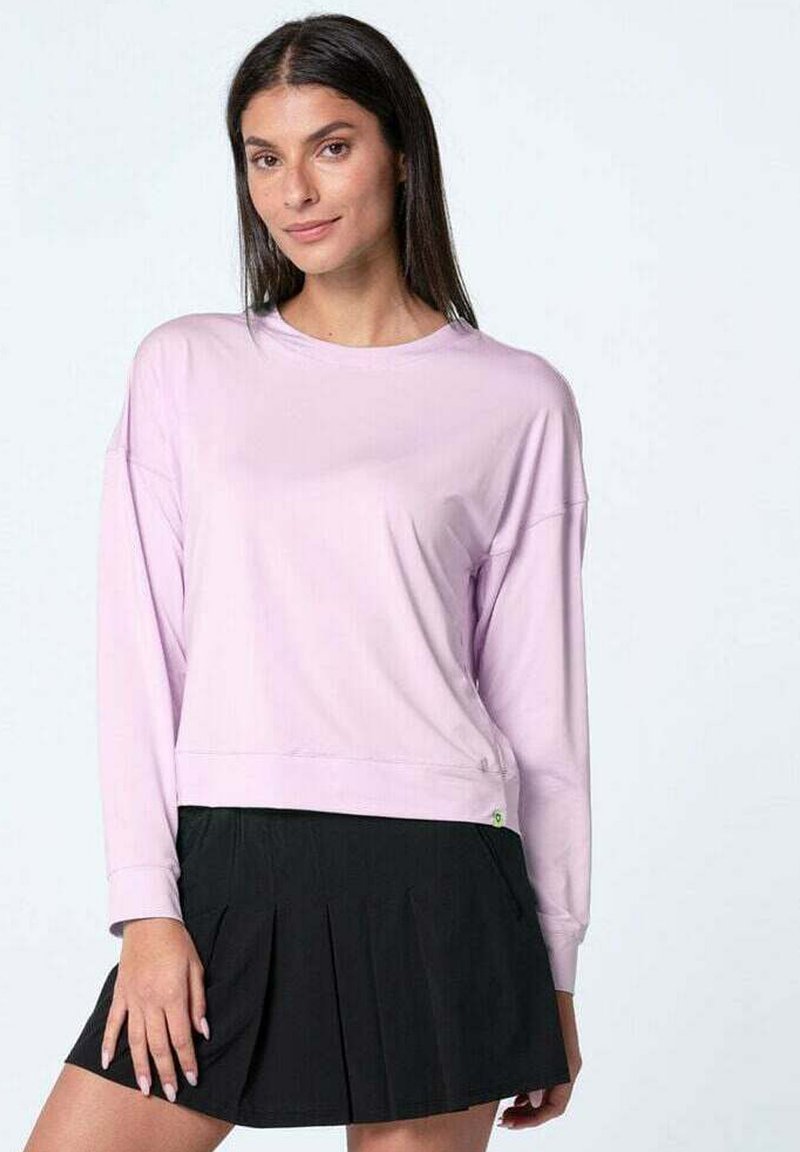 Donna con lunghi capelli scuri che indossa una camicia a maniche lunghe di un leggero colore viola e una gonna nera a pieghe, in piedi contro uno sfondo chiaro e neutro.