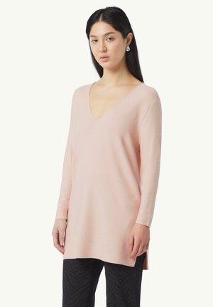 Light pink long-sleeve tunic sweater with a V-neck, ribbed texture, side slits, and a relaxed fit, paired with black textured pants.