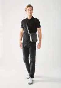 Shoulder bag in black textured material with a front zip pocket, adjustable strap, and a compact rectangular shape. Worn crossbody.