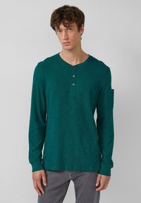 Jonge man met krullend haar draagt een teal lange mouwen henley shirt met knopen en een zak op de bovenste linker mouw, gecombineerd met grijze spijkerbroek.