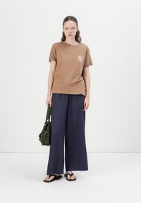 Bruine katoenen T-shirt met een kleine afbeelding op de borst, gecombineerd met losse marineblauwe wijde broek en zwarte flip-flops. Inclusief groene tas.