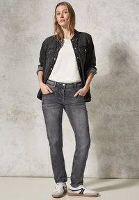 Giubotto di jeans nero sopra una maglietta bianca, abbinato a jeans grigi e sneakers bianche con dettagli neri. Texture liscia, vestibilità casual.