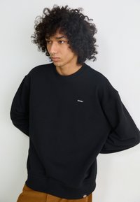 Zwarte sweatshirt met een ronde hals en losse mouwen, voorzien van een klein wit logo label op de voorkant. Zacht materiaal en ribbelboorden.