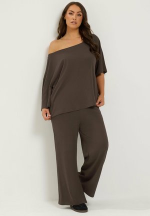 Mujer con blusa de hombros descubiertos de color marrón oscuro y pantalones anchos a juego, de pie frente a una pared blanca.