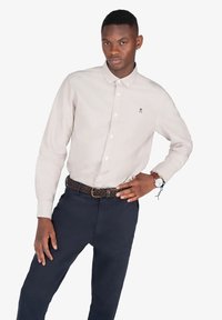 Camisa de botón de manga larga beige claro con cuello y un pequeño logo en el pecho, combinada con pantalones azul marino oscuros y un cinturón trenzado.