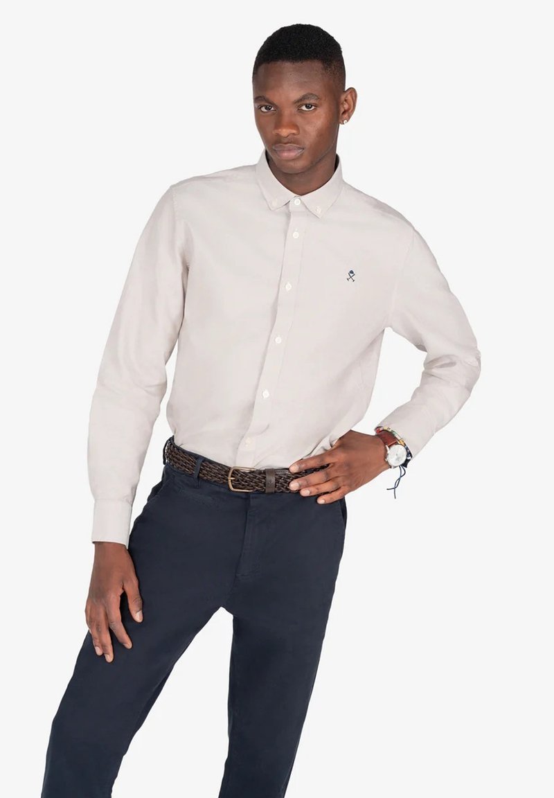 Camisa de botón de manga larga beige claro con cuello y un pequeño logo en el pecho, combinada con pantalones azul marino oscuros y un cinturón trenzado.