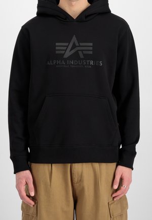 Person iført en sort Alpha Industries-hættetrøje med logo og tekst, kombineret med beige cargobukser, vist fra nakke til hofter.