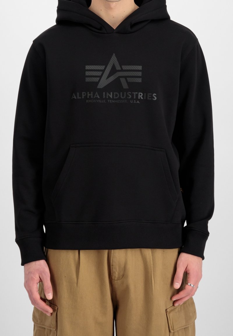 Personne portant un sweat à capuche noir Alpha Industries avec logo et texte, associée à un pantalon cargo beige, visible du cou jusqu'aux hanches.