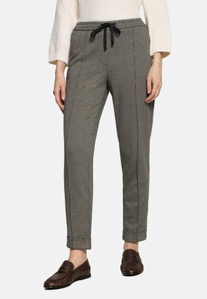 Woman wearing grey checkered tapered pants with black drawstring, white sweater, and brown loafers, standing on white background.
