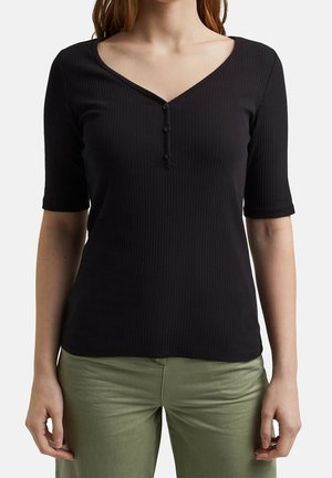 Mujer con una camiseta negra acanalada con medio botones y mangas hasta el codo, y pantalones verde oliva, mostrada desde el cuello hasta las caderas.