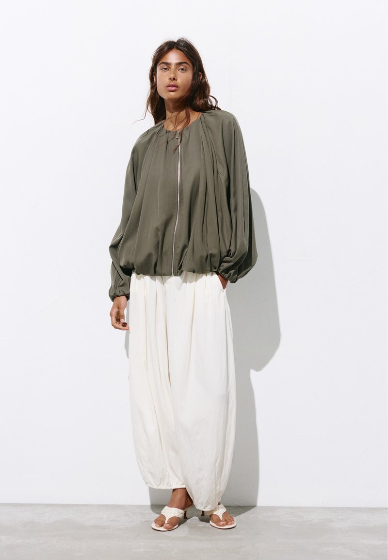 Femme portant une veste zippée ample olive, un pantalon blanc large et des sandales blanches ouvertes à l'avant, debout contre un mur blanc.