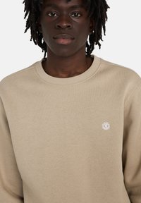 Young person with dark skin and dreadlocks wearing a beige crewneck sweatshirt with a small white circular logo on the chest.