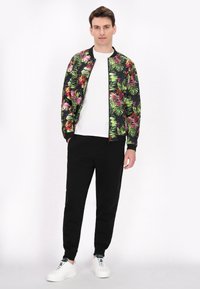 Zwarte bomberjack met tropische bloemenprint in groen, roos en geel. Gecombineerd met zwarte joggers en witte sneakers. Witte t-shirt eronder.