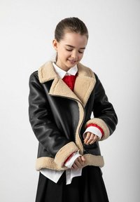 Czarna skórzana kurtka z beżowym futrzanym kołnierzem i mankietami, z zamkiem błyskawicznym i wewnętrzną czerwoną podszewką. Model nosi pod nią białą koszulę.