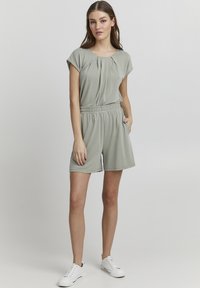 Light green romper with a round neck, short sleeves, pleated front detail, elastic waistband, and side pockets; paired with white sneakers.