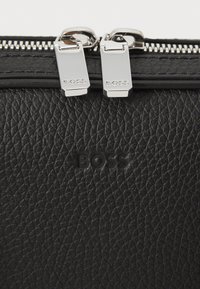 Sort textureret lædertaske med sølvfarvet lynlås og prægede "BOSS" logo. Designet fremhæver funktionalitet og sofistikering.