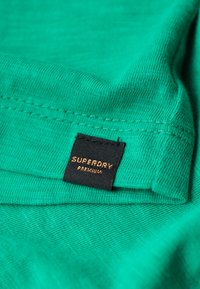 Grøn bomuldsstof med en glat tekstur, der viser et sort mærke med teksten "SUPERDRY PREMIUM" i guldskrift.