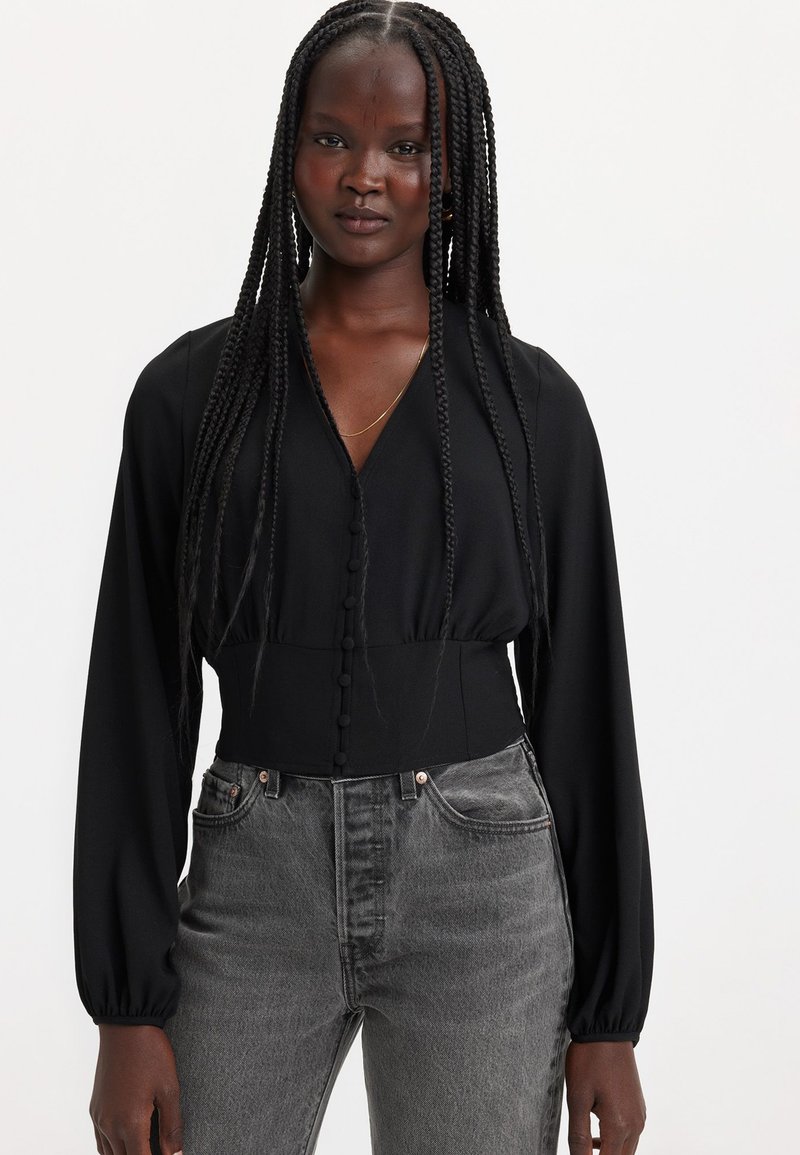 Schwarze Bluse mit langen Ärmeln, V-Ausschnitt, Frontknöpfen und tailliertem Schnitt, kombiniert mit grauen Jeans mit hoher Taille. Glatte Stofftextur.