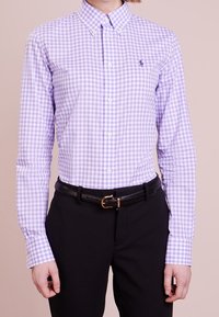 Person som bär en knäppt lila och vit rutig skjorta instoppad i svarta byxor med ett svart bälte, stående mot en enfärgad bakgrund.