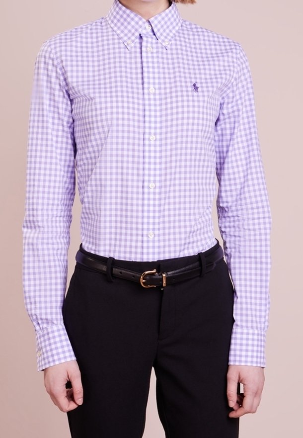 Person som bär en knäppt lila och vit rutig skjorta instoppad i svarta byxor med ett svart bälte, stående mot en enfärgad bakgrund.