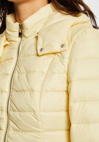 Yellow padded jacket featuring a high collar, snap details on the shoulder, and contrasting stitch lines, with a smooth, shiny texture.
