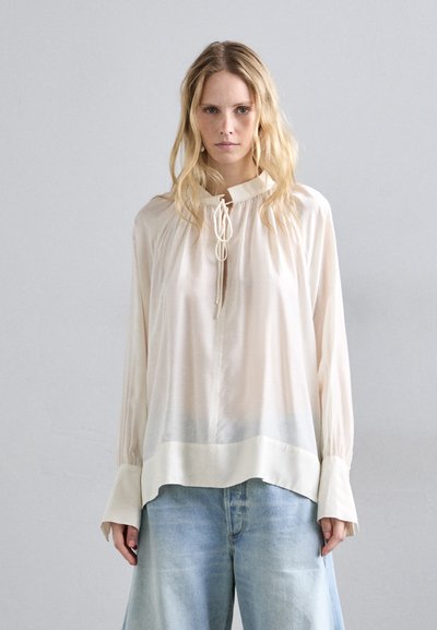 Hellbeige Bluse mit einer Schleifenanfällung, langen Ärmeln und einem leicht lockeren Schnitt. Glatte Textur mit subtiler Ombre-Wirkung am Saum.