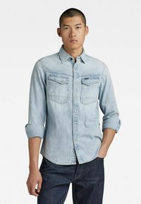 Light blue denim shirt with long sleeves rolled to elbows, featuring two chest pockets and a button-down collar. Smooth texture, classic fit.