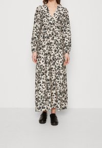 Robe longue florale avec un décolleté en V profond, des manches longues et une base beige. Présente des motifs floraux noirs et verts ; portée avec des sabots noirs.