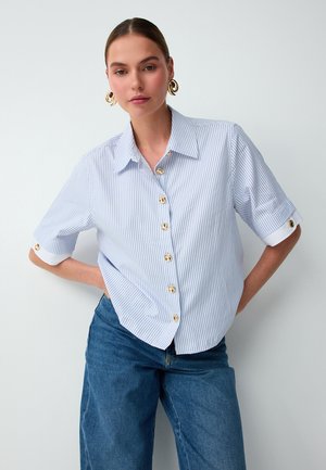 Donna che indossa una camicia a righe azzurro chiaro con bottoni dorati e jeans blu, in piedi con una mano sul fianco su sfondo bianco.