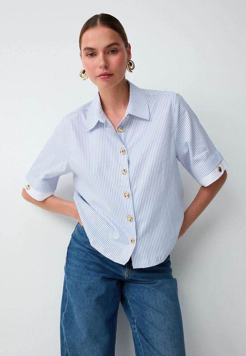 Femme portant une chemise rayée bleu clair à boutons dorés et un jean bleu, debout avec une main sur la hanche devant un fond blanc.