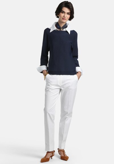 Pulover tricotat bleumarin, purtat peste o cămașă albă cu guler, asortat cu pantaloni albi croiți și pantofi maro toc cu detaliu de fundă.